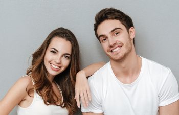 A smiling woman and man, both in white shirts, stand against a light gray wall. The woman has long brown hair and rests her arm on the man's shoulder.