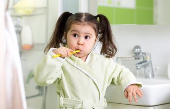 A young child with pigtails is brushing their teeth in a bathroom, wearing a green robe and holding a yellow toothbrush.